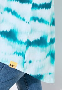 Camiseta azul claro con un patrón de tie-dye en tonos turquoise, con una etiqueta de tela amarilla que muestra un símbolo de infinito en el dobladillo.