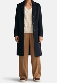 Abrigo largo azul marino con cuello de muesca, cinco botones, sobre un suéter texturizado color crema y pantalones anchos de color tostado, combinados con zapatillas marrones.