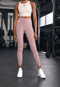 Světle růžové legíny s vysokým pasem vyrobené z elastického materiálu, spárované s přiléhavým bílým sportovním cropped topem s černým logem. Bílé sportovní boty.