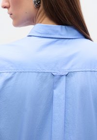 Πλευρικό προφίλ ενός ατόμου με μακριά καστανά μαλλιά που φοράει ασημένια κρίκα σκουλαρίκια και ένα ανοιχτό γαλάζιο πουκάμισο με γιακά και πτυχώσεις στο πίσω μέρος.