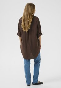 Barna hosszú ing felhajtott ujjal, enyhén magas-alacsony szegéllyel, kék farmerrel és fekete lapos cipővel párosítva, lazább megjelenést mutatva.