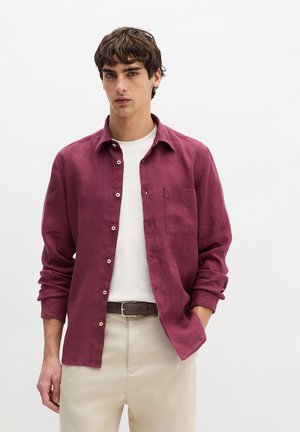 Junger Mann trägt unknöpftes burgunderrotes Langarmhemd über weißem T-Shirt, beiger Hose und dunklem Gürtel vor schlichtem grauen Hintergrund.