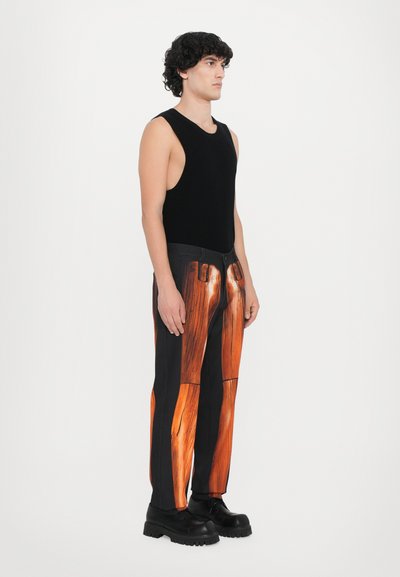 Schwarzes ärmelloses Tanktop, kombiniert mit schwarzen Hosen im Holzmaserungsdesign. Das Model trägt klobige schwarze Schuhe mit dicken Sohlen.