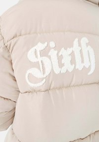 Pufferjack in lichtbeige stof met gestikt wit "Sixth" logo. Beschikt over een gequilte design en een zachte, textuurachtige oppervlakte.