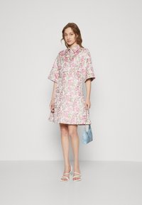 Blumenkleid mit rosa und grünen Mustern, kurzen Ärmeln und einem Kragen. Kombiniert mit einer hellblauen Tasche und weißen Sandalen mit Riemchen.