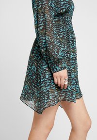 Abito fluido e stampato con un design a squame verde e blu, tessuto trasparente, maniche lunghe e vita arricciata con un orlo leggermente asimmetrico.