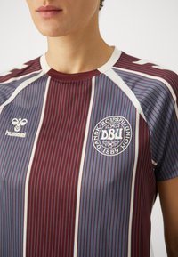 Bordo ja sinise triibuline spordimüts valgete aktsentidega. Omab ümmargust kraed ja ümmargust logo vasakul rinnal. Valmistatud hingavast kangast.