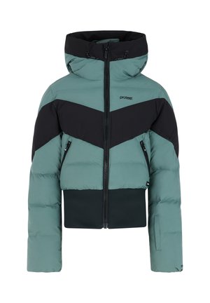 Jonge persoon met zwarte muts, blauwgroene en zwarte pufferjas, zwarte broek en zwarte laarzen staand tegen een witte achtergrond.