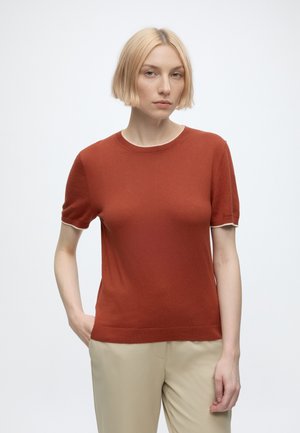 Femme blonde portant un pull à manches courtes couleur rouille et un pantalon beige, debout avec une main dans la poche sur un fond uni.