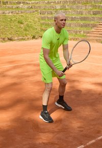 Conjunto deportivo verde neón de manga corta y pantalones cortos, zapatillas deportivas negras, sosteniendo una raqueta de tenis en una cancha de tierra batida.