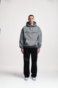Gray hooded sweatshirt with "2Y STUDIOS" printed in black, paired with black ripped jeans and light blue sneakers. Plain background.