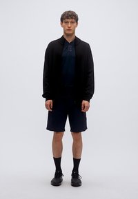 Μαύρο μπουφάν με φερμουάρ, ναυτικό μπλουζάκι με κοντά μανίκια και σκούρα σορτς. Το μοντέλο φορά μαύρα παπούτσια και κάλτσες, στέκεται μπροστά σε απλό λευκό φόντο.