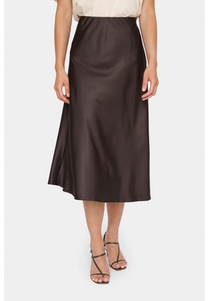 Vrouw draagt een donkerbruine satijnen midirok met hoge taille en een split aan de zijkant, gecombineerd met een beige top en sandalen met hoge hakken en bandjes.