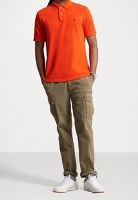 Pomarańczowa koszulka polo z niebieskim logo, w połączeniu z jasnobrązowymi spodniami cargo z bocznymi kieszeniami i luźnym krojem. Białe sneakersy.