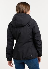 Donna che indossa una giacca nera con cappuccio e jeans blu, voltata di spalle, con una mano rilassata al suo fianco su uno sfondo semplice.