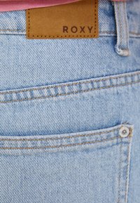 Light blue denim shorts with a brown leather Roxy label. Features minimalistic stitching and a button detail on the back pocket.