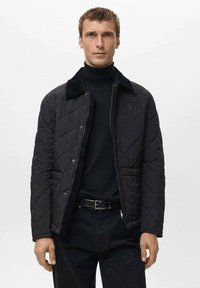 Giacca nera trapuntata con collo peloso, bottoni a pressione frontali e due tasche laterali. Indossata sopra un dolcevita nero e pantaloni neri.