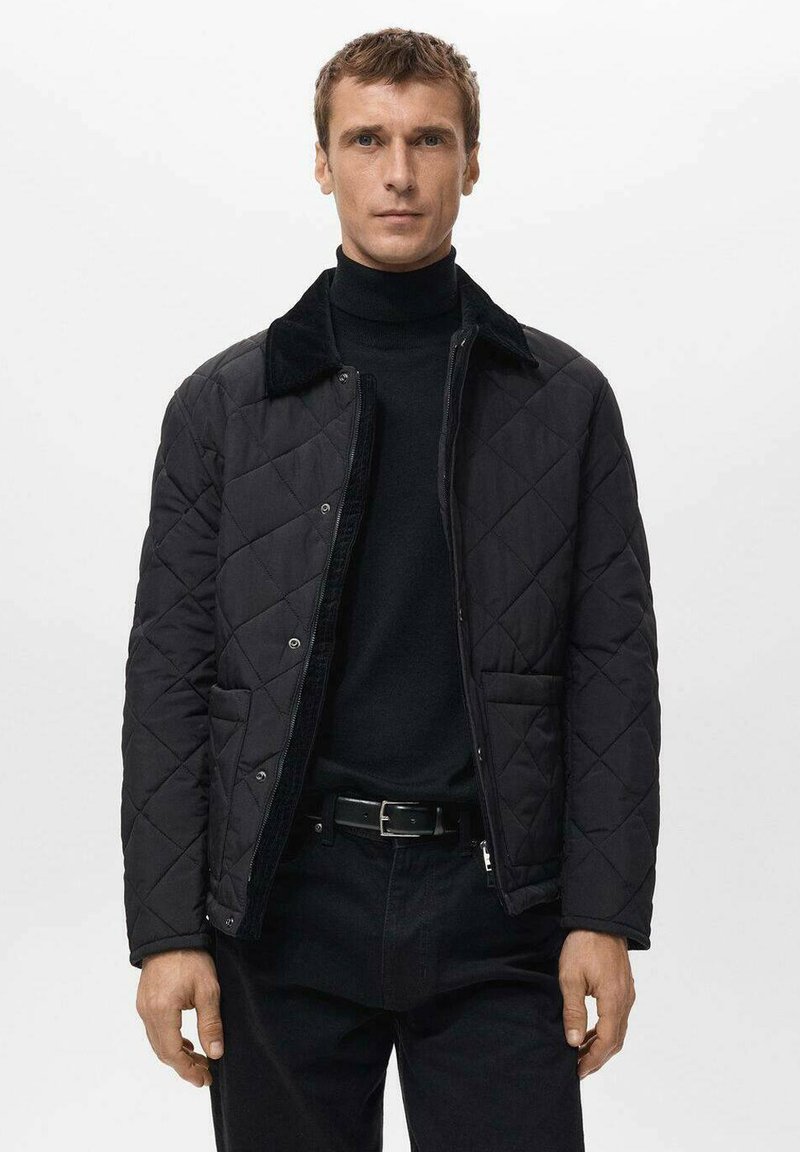 Giacca nera trapuntata con collo peloso, bottoni a pressione frontali e due tasche laterali. Indossata sopra un dolcevita nero e pantaloni neri.