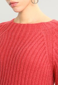 Pull en maille corail vif, avec une texture côtelée et un col large. Matière douce et légère qui améliore le confort et la portabilité.
