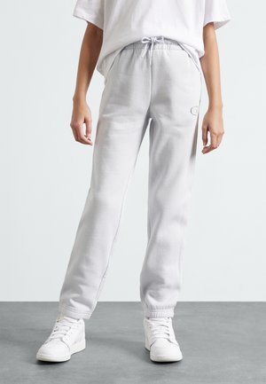 Light gray sweatpants in soft fabric with a drawstring waistband, cuffed ankles, and a small logo detail on the side. Styled with white sneakers.