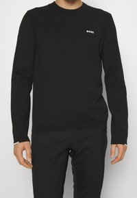 Pull noir à col rond en matière tricotée. Présente un logo de marque subtil sur la poitrine, des poignets côtelés et un détail aux épaules. Silhouette ajustée.