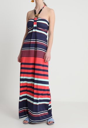 Femme portant une robe longue bustier rayée multicolore avec un collier de perles et des sandales bleu clair à bouts ouverts sur un fond uni.