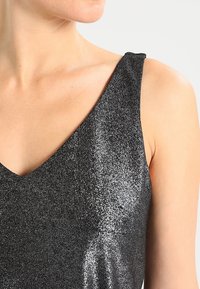 Schwarzes ärmelloses Oberteil mit V-Ausschnitt. Hergestellt aus einem glänzenden, strukturierten Stoff mit funkelndem Finish, der Konturen betont und Licht reflektiert.