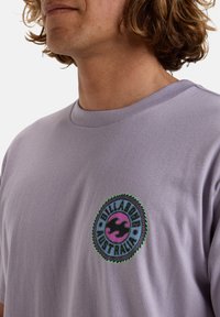 Camiseta de un ligero color púrpura con un logotipo bordado redondo. El logotipo presenta un diseño de ola negra y el texto "BILLABONG AUSTRALIA" en un patrón circular.