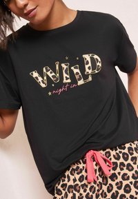 Černé tričko s krátkým rukávem, na kterém je vytištěno "WILD" s leopardím vzorem a růžovými akcenty. Kombinováno s béžovými kalhotami zdobenými leopardími skvrnami a růžovým stahovacím šňůrkou.