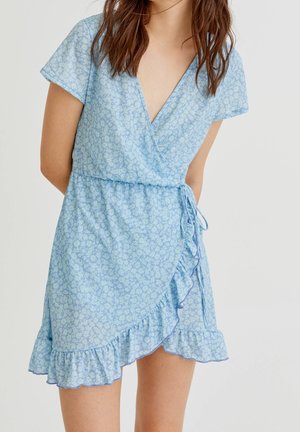 Kvinde iført en kortærmet, lyseblå blomsterprintet slå-om-kjole med flæsekant og bindebånd i siden, stående mod en ensfarvet baggrund.