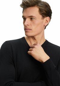 Schwarzer langärmliger V-Ausschnitt-Pullover mit gerippter Textur. Das Modell posiert mit der Hand am Kragen und präsentiert das figurbetonte Design und den glatten Stoff.