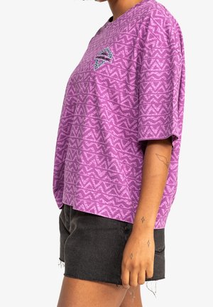 Osoba ubrana w luźną fioletową koszulę z wzorem oraz czarne szorty jeansowe, pokazująca tatuaże na przedramieniu i nadgarstku.