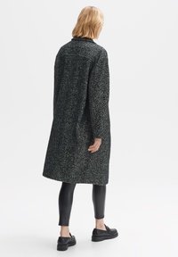 Long black and white herringbone coat with a collar, side pockets, and a relaxed fit. Worn over black leggings and flat shoes.