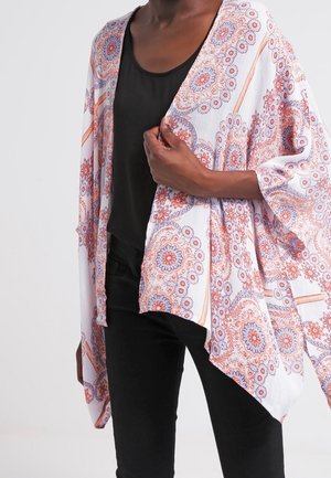 Osoba nosząca luźne białe kimono z różowymi, pomarańczowymi i niebieskimi kwiatowymi wzorami na czarnym topie i czarnych spodniach, trzymająca kimono zamknięte.