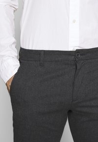 Muž v bílé košili se zapínáním na knoflíky, zastrčené v tmavě šedých kalhotách s proužky, s rukou v kapse na šedém pozadí.