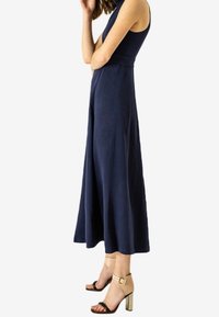Námořnická modrá bezramínková šaty s upnutým živůtkem a rozšířenou sukní. Modelka má na sobě béžové sandály na podpatku s řemínkem kolem kotníku a obdélníkovou sponou.