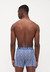Caleçons pour hommes présentant un motif floral bleu sur une base blanche, avec une taille bleue unie marquée "BJÖRN BORG". Texture de tissu lisse.