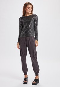 Top lungo a maniche di raso nero con paillettes, scollatura rotonda, abbinato a pantaloni cargo grigi morbidi e sandali alti neri.