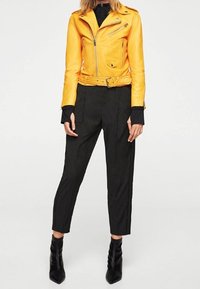 Žlutá kožená motocyklová bunda se zipovými detaily, v kombinaci s černými kalhotami s vysokým pasem a černými kotníkovými botami. Hladká textura a přiléhavý design.