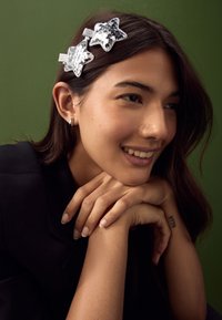 Silberne haarnadeln in sternform mit Pailletten und glatter Oberfläche, getragen von einer Person mit langen, dunklen Haaren vor einem grünen Hintergrund.