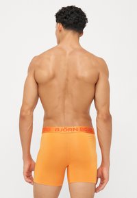 Boxers de tom laranja com ajuste justo, apresentando uma faixa vermelha com a inscrição "BJÖRN BORG" em letras grandes. Textura suave, design minimalista.