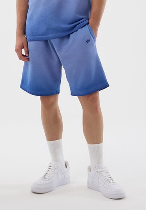 Person iført blå gradientshorts, hvide crew-strømper og hvide sneakers, stående mod en ensfarvet baggrund.