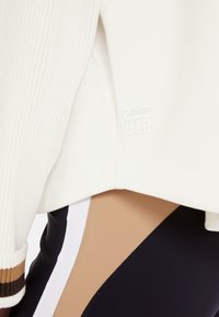 White ribbed sweater with a cropped design, featuring a subtle logo in white. Accompanied by fitted leggings with color-blocking in beige and navy.