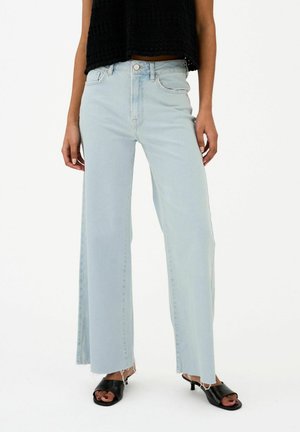 Persoon die lichtblauwe jeans met brede pijpen en onafgewerkte zomen draagt, gecombineerd met zwarte sandalen met hak en open teen en een zwart mouwloos topje.