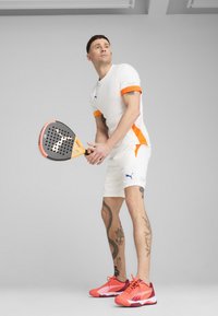 Camiseta deportiva blanca con acentos naranjas, pantalones cortos blancos y zapatos en un vibrante tono rosa-naranja. Sosteniendo una pala negra con detalles naranjas.