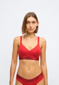 Rød bomulls-bralette med tynne stropper og V-hals-design. Har en kontrasterende svart 'Calvin Klein'-logo på elastikkbåndet.