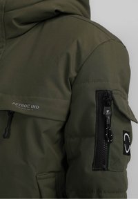 Manche de veste matelassée vert foncé avec poche zippée noire, languette en tissu et petit écusson logo noir sur le haut du bras.
