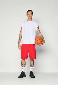White sleeveless top with black Nike logo, red shorts, black sneakers, and white socks holding a basketball. Tattoos visible on arms.