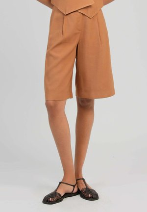 Jambes portant un short marron ample mi-longueur associé à des sandales plates tressées à bride en T marron foncé sur fond neutre.