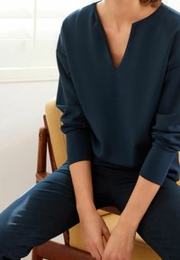 Haut bleu marine à manches longues avec un col en V, texture lisse et coupe décontractée. Porté avec un pantalon assorti bleu marine, assis sur une chaise en bois.